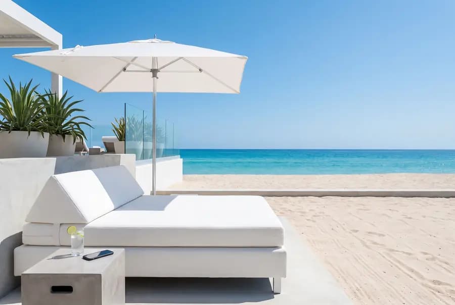 A white daybed under a white umbrella at a beach club, a drink and a phone on a side table, pale sand and a turquoise sea.