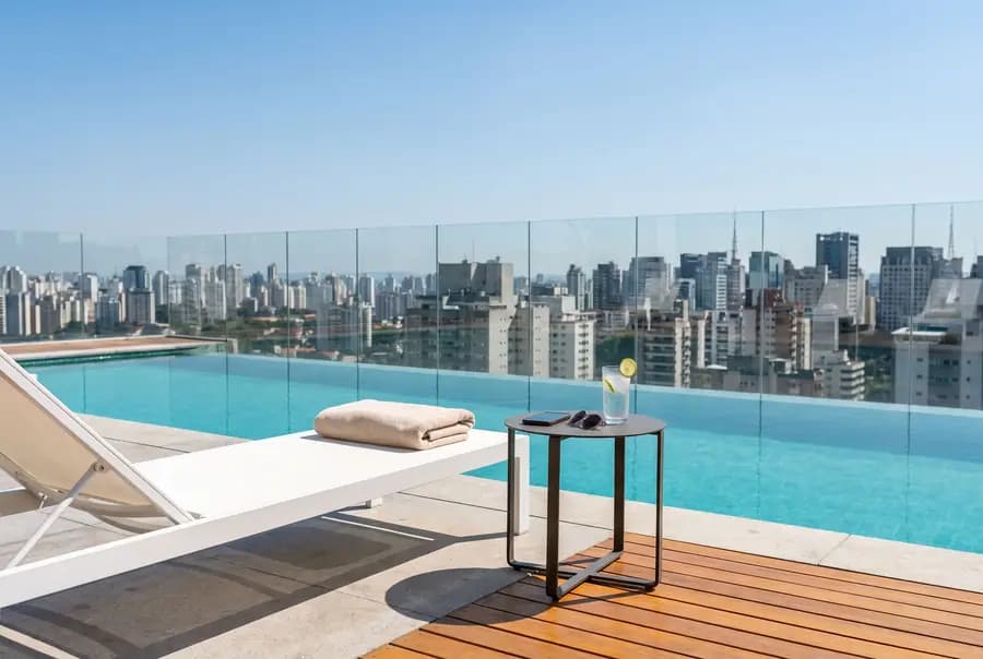 A white lounge chair with a folded towel beside a rooftop pool, a side table with a drink and a phone, and a city skyline under a blue sky.